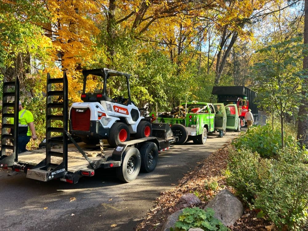 Construction vehicles and equipment on a trailer in a colorful autumn forest setting.