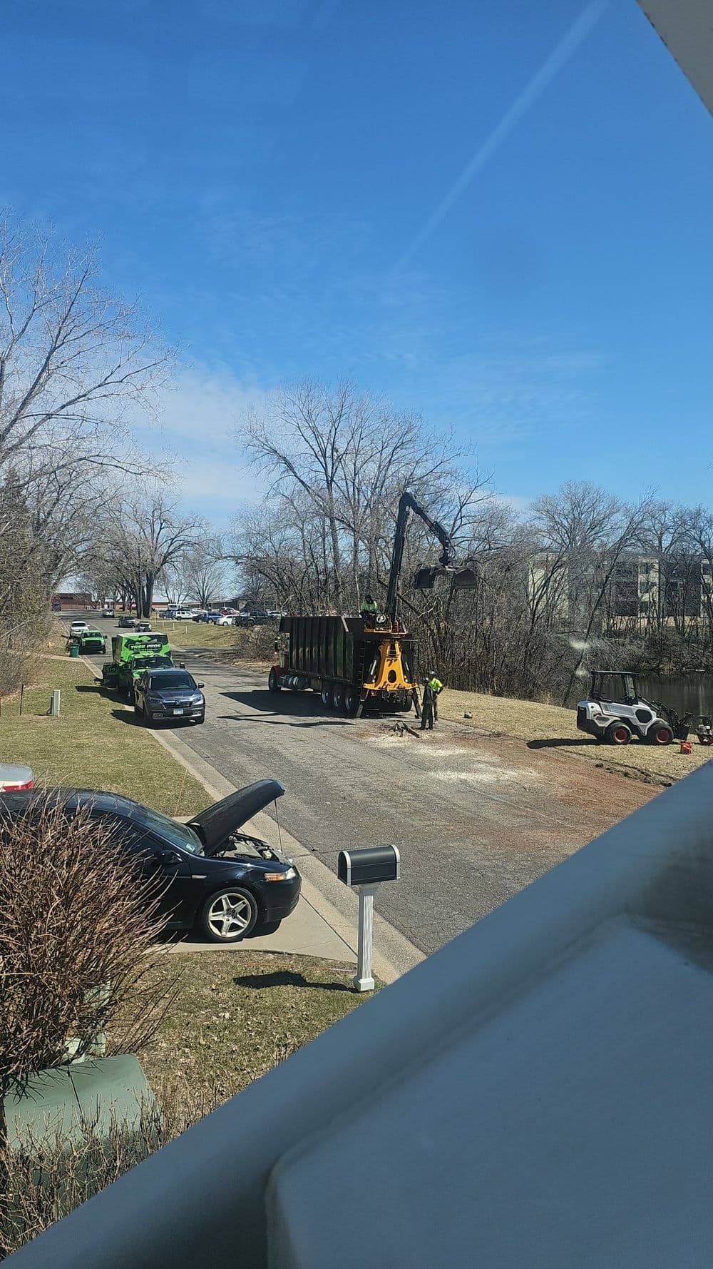 Worker using a crane to load debris into a truck on a sunny day near a riverbank.