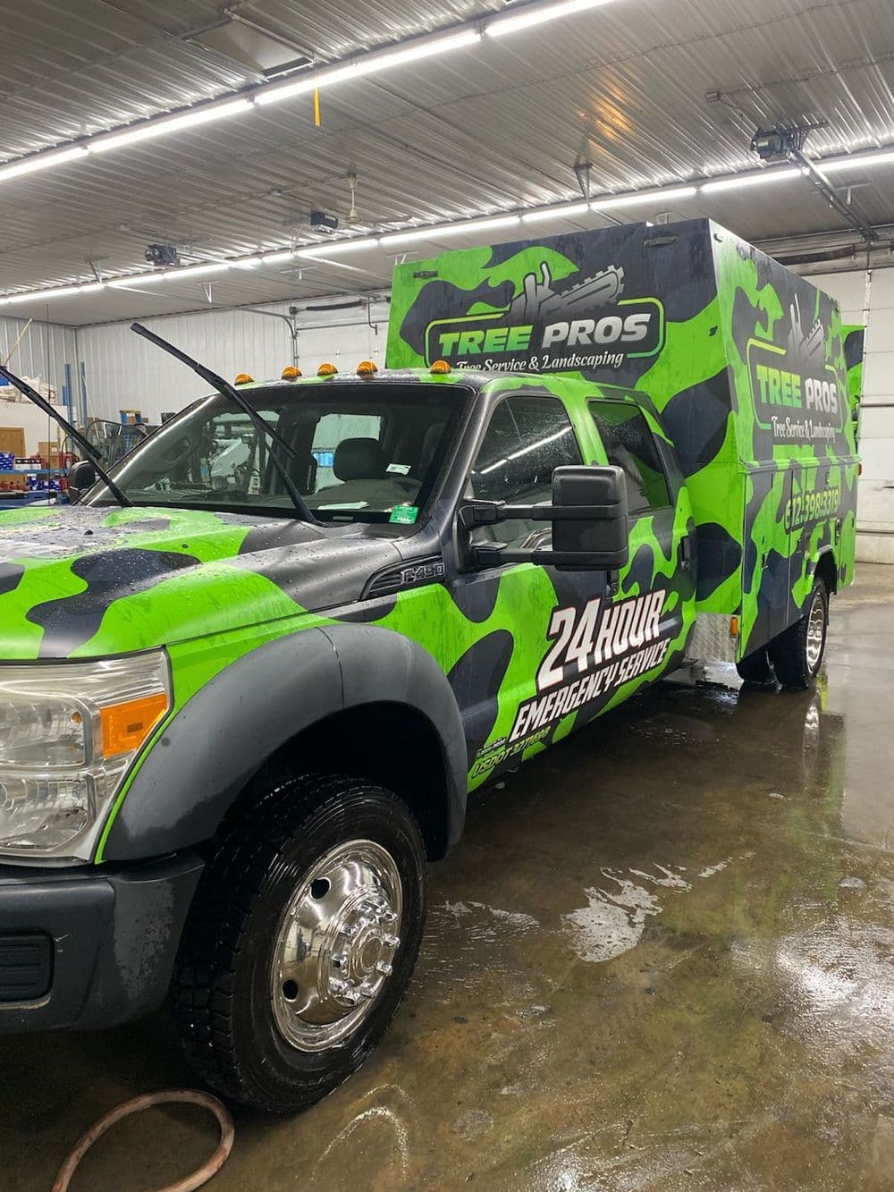 Green and black Tree Pros truck marked for 24-hour emergency service, parked indoors.
